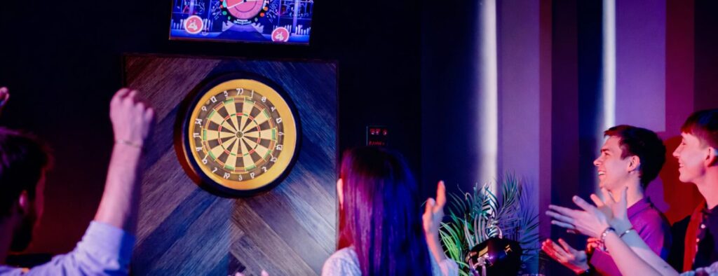 Friends playing interactive darts in a neon-lit sports bar with a digital scoreboard.